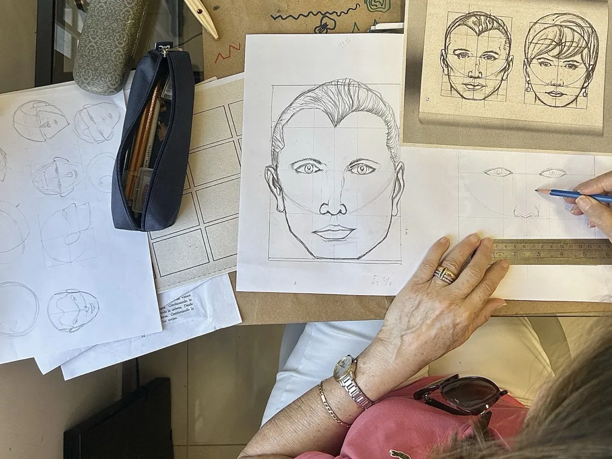 Mãos desenhando as proporções de um rosto a lápis, vistas de cima
